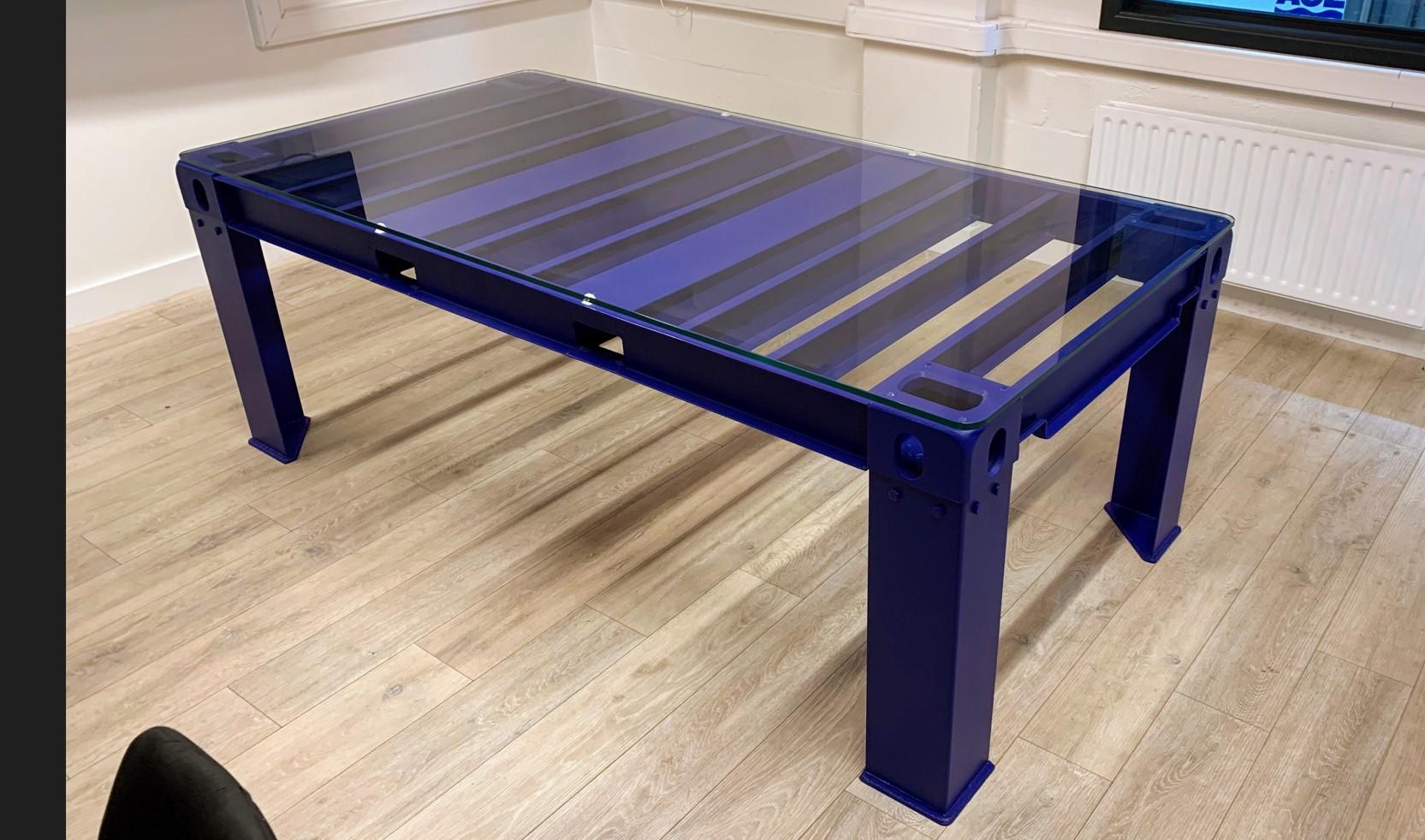 Custom table made from spare shipping container materials in dark blue with glass top and four legs in office room 